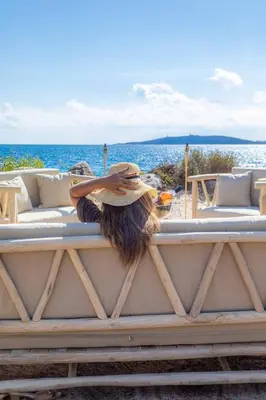 a woman with a hat sitting on a bench near the ocean at Villa Cala Rossa pieds dans l'eau Porto-Vecchio in Lecci