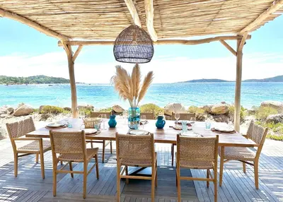 a dining table and chairs on a deck with the ocean at Villa Cala Rossa pieds dans l'eau Porto-Vecchio in Lecci